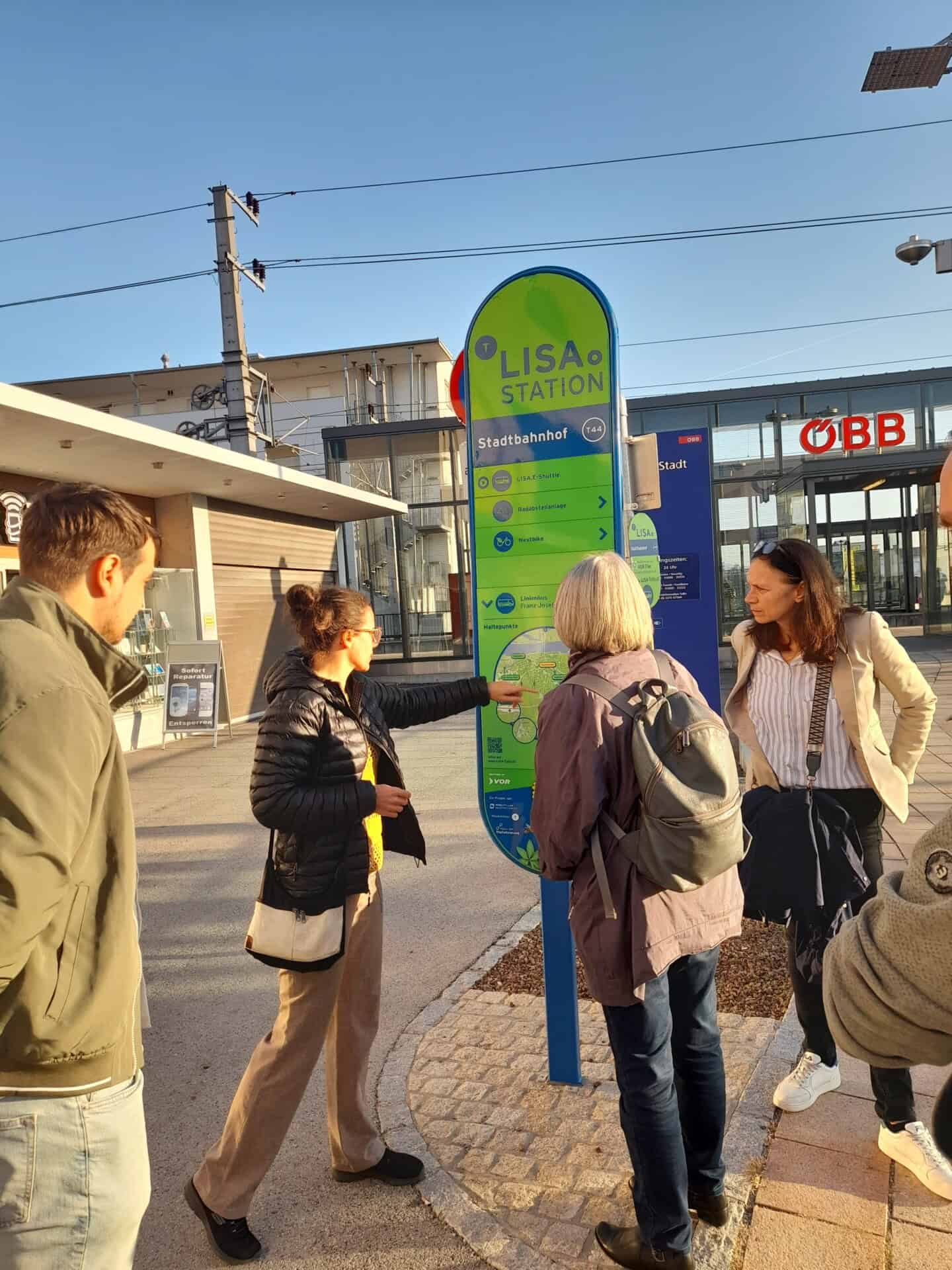 Personen stehen vor Bahnhof und sehen sich Infostele an