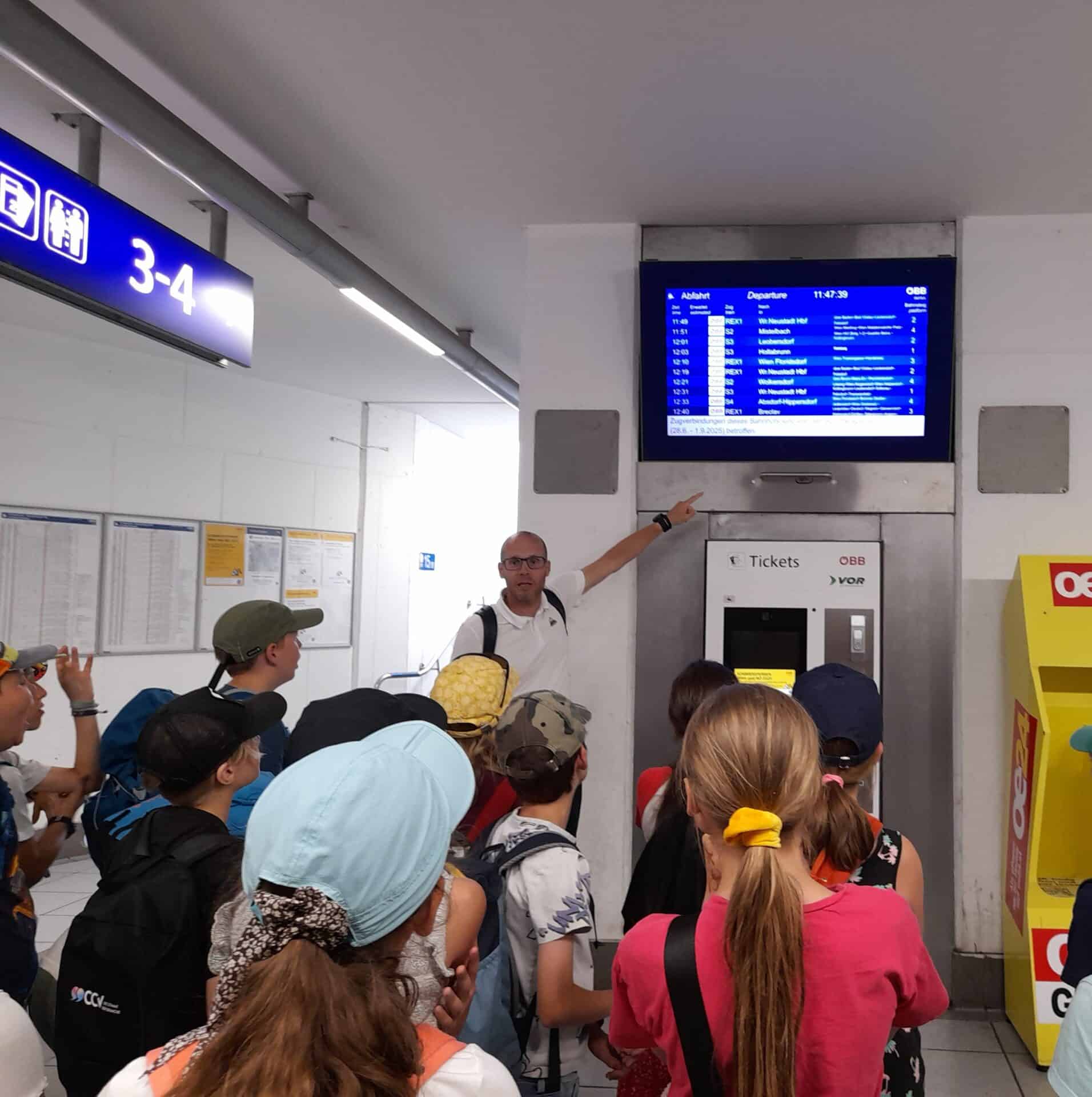 Mann erklärt Kinder die Anzeigetafel am Bahnhof Mödling