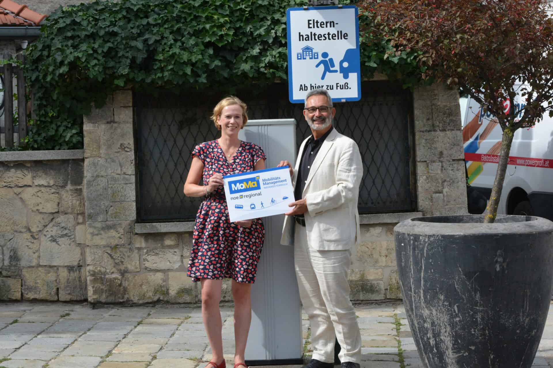 Verkehrsberaterin und Vizebürgermeister stehen mit Fototafel vor einem Elternhaltestellenschild