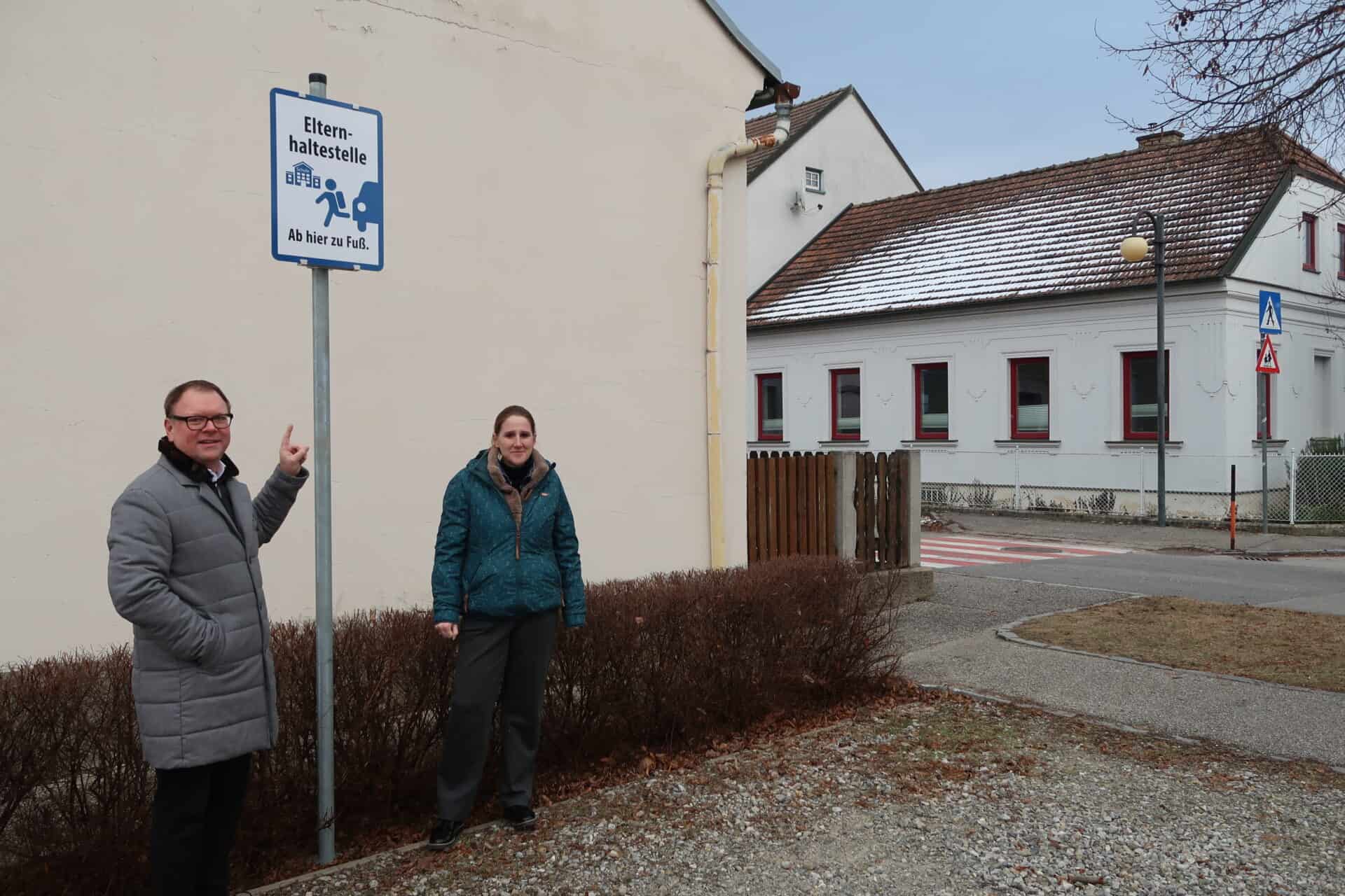 Zwei Menschen stehen auf einem Parkplatz, der als Elternhaltestelle gekennzeichnet ist.