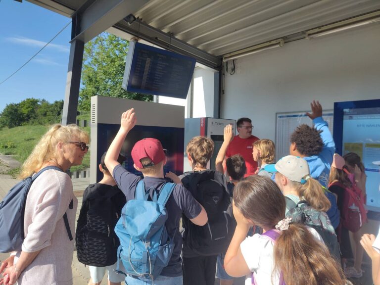 Kinder stehen an Bahnhaltestelle und lernen über die Anzeigetafel